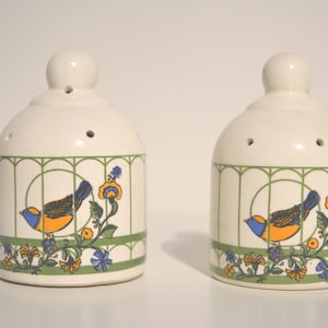 May include: Two white ceramic salt and pepper shakers with a bird and floral design. The shakers have a green, white, and yellow design with a blue bird on each.