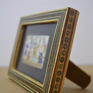 May include: A small, rectangular picture frame with a detailed, multi-colored design. The frame features a black interior, a decorative border, and a wooden stand. The artwork inside depicts figures in blue, red, and yellow.