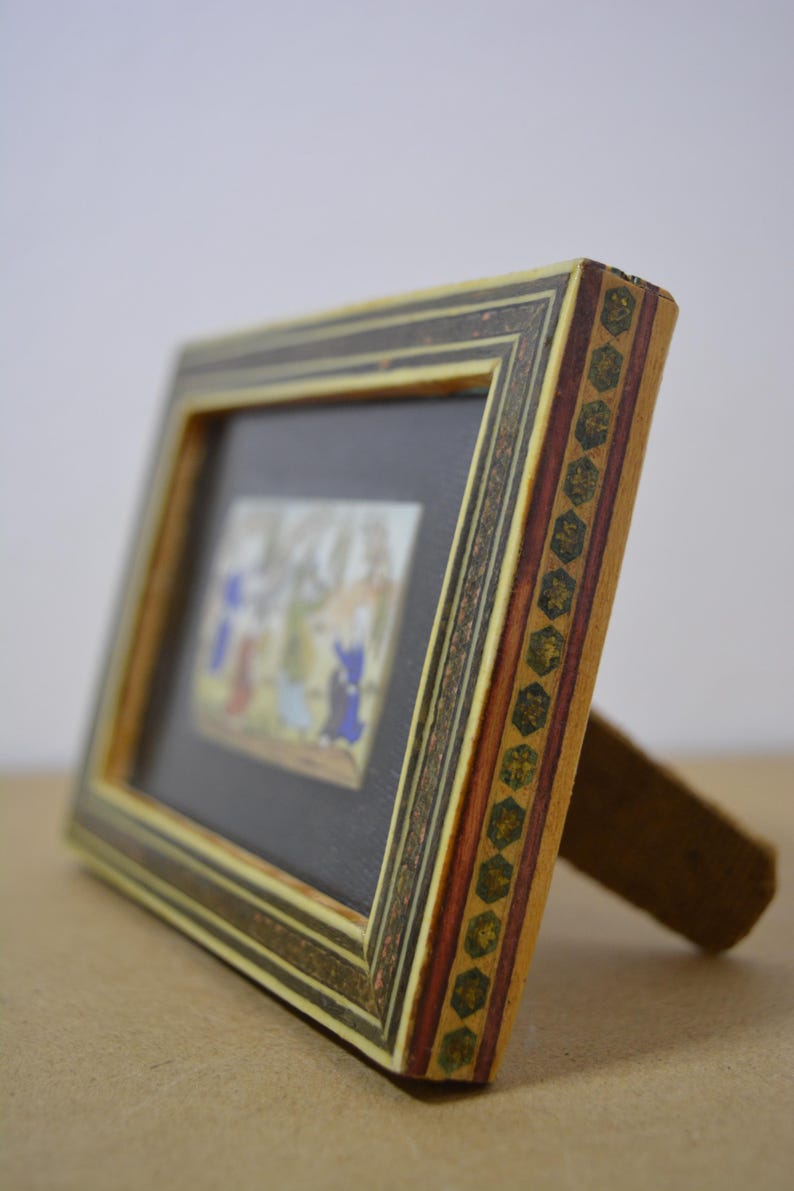 May include: A small, rectangular picture frame with a detailed, multi-colored design. The frame features a black interior, a decorative border, and a wooden stand. The artwork inside depicts figures in blue, red, and yellow.