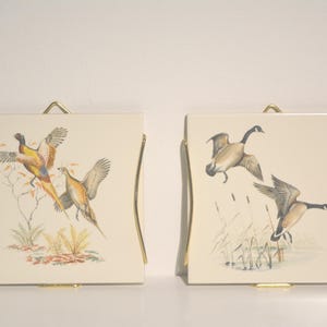 May include: Two ceramic tiles with gold metal frames, each featuring a different bird illustration. The first tile shows two pheasants in flight, while the second tile depicts two geese flying over a marsh.