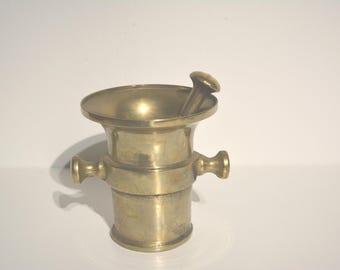 May include: A brass mortar and pestle with a round bowl and a handle on each side. The pestle is attached to the bowl.