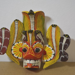 Op de afbeelding: Een handgeschilderd houten masker met een centraal rood gezicht, een brede grijns en grote ogen. Het masker is versierd met gele en bruine cobra-achtige vormen, wat een gedetailleerd ontwerp creëert. Het masker is waarschijnlijk voor decoratieve doeleinden.