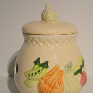 Pode incluir: Pote de cerâmica creme com tampa e desenhos de vegetais. O pote apresenta ilustrações em relevo de ervilhas, uma cebola e outros vegetais. A tampa tem um botão decorativo. O pote tem um padrão tecido na parte superior.