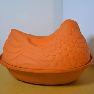 May include: A terracotta-colored chicken-shaped ceramic baker. The baker has a detailed design, with a rounded base and a lid shaped like a chicken. The lid features a detailed head, body, and tail feathers. The baker is designed for cooking.