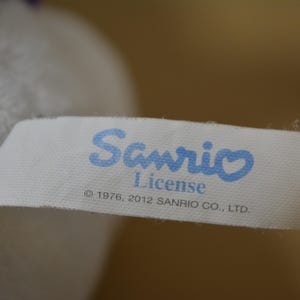 May include: A white fabric tag with the blue text "Sanrio License" and the copyright information "&copy; 1976, 2012 SANRIO CO., LTD." is attached to a plush toy. The tag has a light blue heart over the letter "i" in Sanrio.