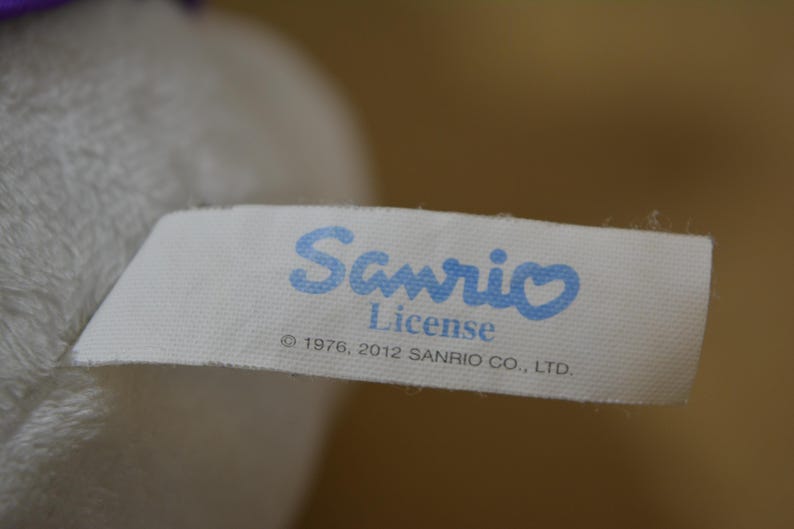 May include: A white fabric tag with the blue text "Sanrio License" and the copyright information "&copy; 1976, 2012 SANRIO CO., LTD." is attached to a plush toy. The tag has a light blue heart over the letter "i" in Sanrio.