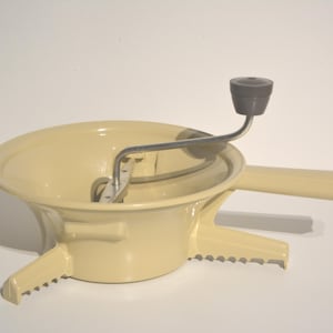 May include: A cream-coloured food mill with a silver crank and a grey handle. The mill has a bowl with a strainer and a base with three legs.