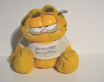 May include: A yellow and orange plush toy of Garfield the cat wearing a white shirt with the text "I'm not FAT I'm underTALL".