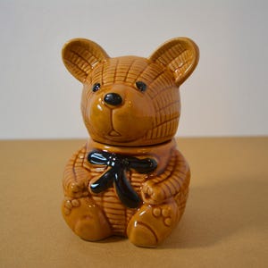 May include: A brown ceramic teddy bear cookie jar with a black bow tie. The bear has a glossy finish and detailed texture, with visible ears, paws, and facial features. The jar is designed to resemble a classic teddy bear.