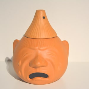 Pode incluir: Um queimador de incenso de cerâmica de terracota em forma de rosto chorando com uma tampa em forma de cone. O queimador é de cor laranja claro e tem um pequeno furo no topo da tampa.