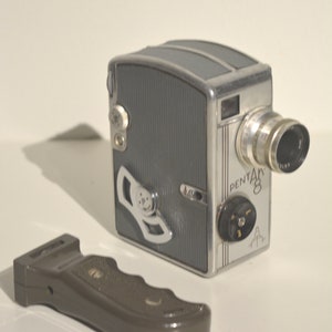 Puede incluir: Una cámara de cine vintage Pentax 8 plateada y gris con una lente negra y un mango de plástico marrón. La cámara tiene un botón plateado en el lateral y el texto "PENTAX 8" impreso en el lateral.