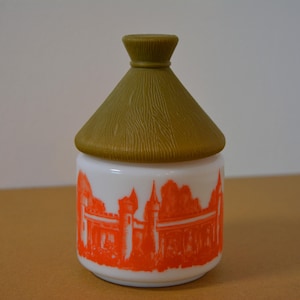 May include: A white jar with an orange castle design, topped with a textured, olive-green lid. The jar's design features a detailed illustration of a castle or palace. The lid has a conical shape.