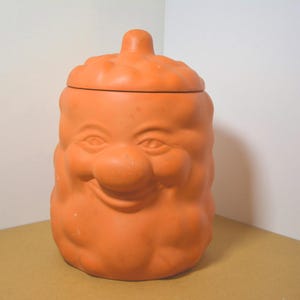 Può includere: Un barattolo per biscotti color terracotta con un design a forma di viso sorridente. Il barattolo ha un coperchio e un corpo arrotondato, con tratti del viso tra cui occhi, naso e bocca sorridente. Il barattolo è in ceramica.