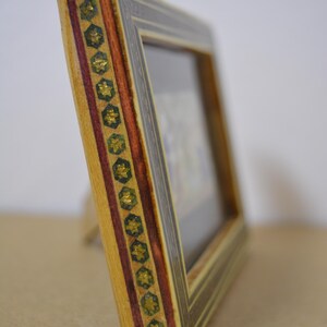 May include: A decorative wooden picture frame with a detailed design. The frame features a light wood base, accented with dark brown and red stripes, and gold star-shaped embellishments. The frame is angled, showcasing its intricate craftsmanship.
