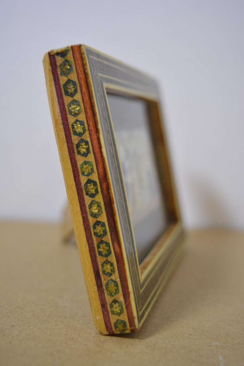 May include: A decorative wooden picture frame with a detailed design. The frame features a light wood base, accented with dark brown and red stripes, and gold star-shaped embellishments. The frame is angled, showcasing its intricate craftsmanship.