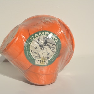 Peut inclure: Un ensemble de gobelets de camping en plastique orange dans un emballage en plastique transparent. Les gobelets sont empilés et portent une étiquette verte sur laquelle est inscrit "Camping" avec une image d'une femme en tenue de camping. L'étiquette indique également "DBP Plastics, Inc."