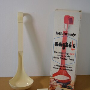 Könnte beinhalten: Eine cremefarbene Vintage-Löffelwaage mit passendem Griff und einem kleinen, separaten Teil. Auf der daneben stehenden Box steht "löffelwaage weighde" und "the amazing new ladle-scale from Switzerland". Die Box hat auch eine rote Schöpfkelle-Grafik.