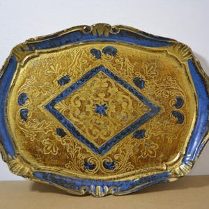 Op de afbeelding: Oud, sierlijk dienblad met een goud- en blauw kleurenschema. Het dienblad heeft een gouden oppervlak met ingewikkelde bloempatronen en een centraal blauw ruitvormig ontwerp. De randen zijn blauw en het heeft decoratieve handvatten.