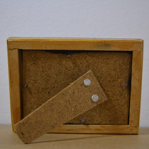 May include: A rectangular wooden picture frame, viewed from the back, with a light brown finish. The frame has a brown backing and a hinged stand. Two small silver magnets secure the stand.