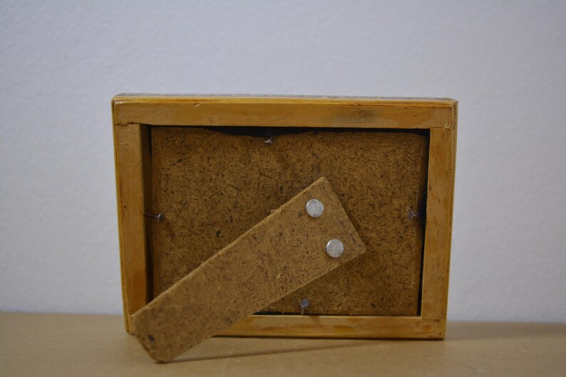 May include: A rectangular wooden picture frame, viewed from the back, with a light brown finish. The frame has a brown backing and a hinged stand. Two small silver magnets secure the stand.