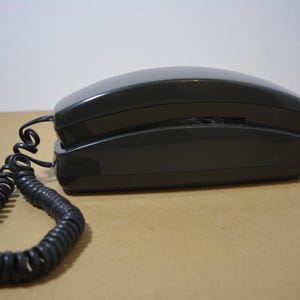 Può includere: Un telefono grigio scuro in stile vintage con un cavo a spirale per il ricevitore. Il telefono ha un design elegante e arrotondato ed è appoggiato su una superficie marrone chiaro. Il cavo a spirale si estende dal ricevitore alla base del telefono.