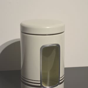 Puede incluir: Un tarro de cerámica blanco con una franja marrón y una ventana transparente. El tarro tiene una tapa.