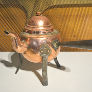 Könnte beinhalten: Eine Kupferteekanne mit einem abgerundeten Deckel und einem langen Ausguss. Die Teekanne hat drei Beine und steht auf einer weißen Oberfläche.