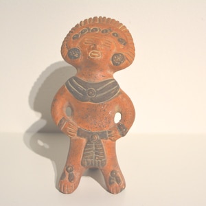 Op de afbeelding: Een terracotta-kleurig beeldje van een staande figuur met armen langs de zijkanten. De figuur heeft een hoofdtooi, een halsketting en een patroonriem. Het beeldje is waarschijnlijk een representatie van een oude godheid of culturele figuur.