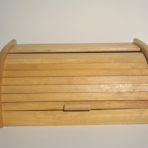 Könnte beinhalten: Eine Brotbox aus Holz mit einem Rolldeckel. Der Deckel besteht aus dünnen Holzlatten und hat einen kleinen Holzgriff.