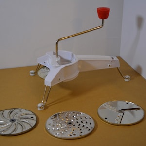 May include: A white, vintage food mill with a red handle and three interchangeable stainless steel blades. The mill has a long arm and is supported by three legs. The blades have different cutting patterns.