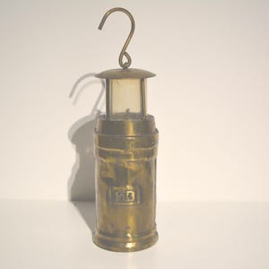 Op de afbeelding: Een hangende messinglamp met een heldere glazen kap en een haak aan de bovenkant. De lamp heeft de letters "R.A." gegraveerd aan de zijkant.