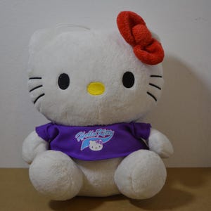 May include: A plush Hello Kitty doll with a white body, black eyes, and whiskers. It has a red bow and wears a purple shirt with the Hello Kitty logo. The doll is sitting on a brown surface.