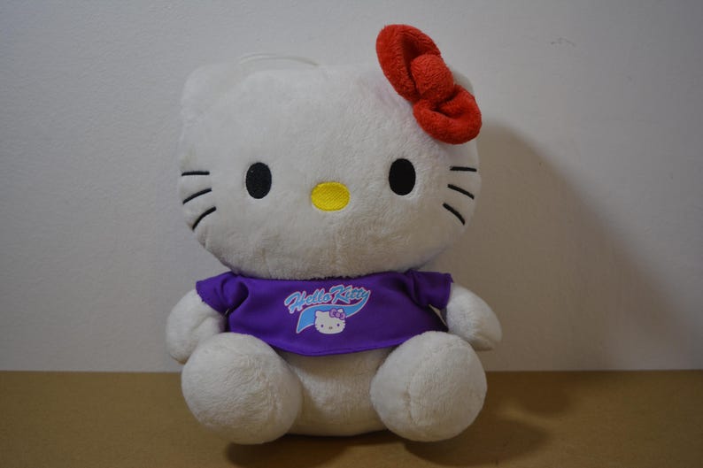 May include: A plush Hello Kitty doll with a white body, black eyes, and whiskers. It has a red bow and wears a purple shirt with the Hello Kitty logo. The doll is sitting on a brown surface.