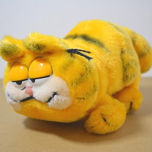 Puede incluir: Un peluche de Garfield amarillo en posición acostada. El peluche tiene rayas y detalles negros, ojos grandes de color naranja, una nariz rosa y un hocico blanco. El juguete está sobre una superficie de tono neutro.
