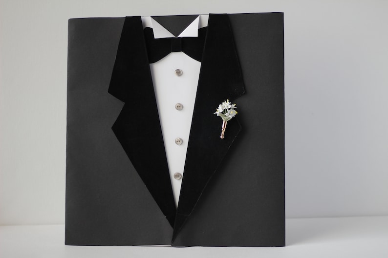 May include: A black tuxedo jacket with a white shirt and a black bow tie. A small white flower boutonniere is pinned to the lapel.