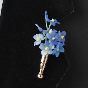 Könnte beinhalten: Ein kleiner Strauß blauer Vergissmeinnicht-Blumen mit gelben Zentren, befestigt an einem goldfarbenen Stiel.