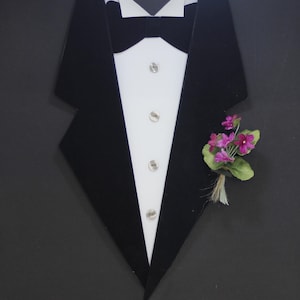 Può includere: Una giacca da smoking nera con camicia bianca e papillon nero. Un piccolo bouquet di fiori rosa è appuntato sul risvolto.