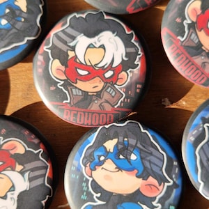 Może przedstawiać: Kolekcja okrągłych, czarnych i czerwonych przypinek z postaciami Red Hood i Nightwing. Przypinki Red Hood przedstawiają postać z czerwoną maską i napisem "Red Hood". Przypinki Nightwing przedstawiają postać z niebieską maską i napisem "Nightwing".