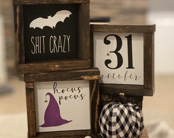 May include: Three square wooden signs with dark brown frames. One black sign says SHIT CRAZY with a white bat. One white sign says 31 OCTOBER with a spider. One white sign says hocus pocus with a purple hat. A black and white plaid pumpkin and a bead garland.