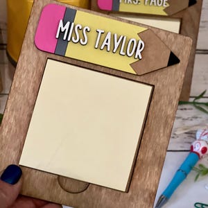 May include: A wooden pencil-shaped frame with a yellow and brown pencil design and a sticky note pad. The frame has the text "Miss Taylor" on it.