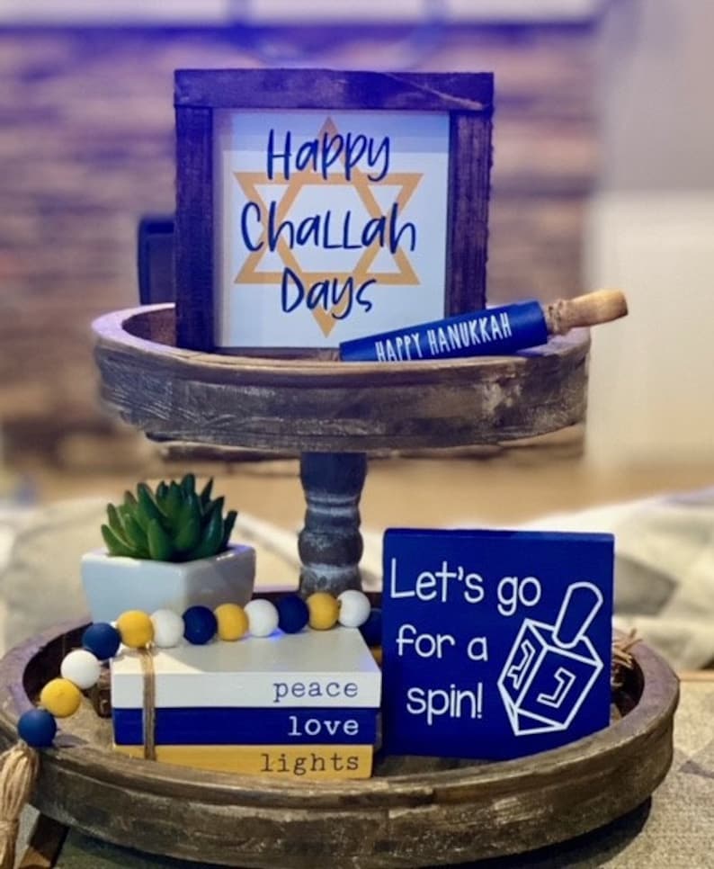 K&ouml;nnte beinhalten: Ein zweist&ouml;ckiges Holztablett, verziert mit blauen und gelben Akzenten. Die obere Ebene zeigt ein Schild mit der Aufschrift "Happy Challah Days" mit einem gelben Davidstern. Die untere Ebene zeigt ein Schild mit der Aufschrift "Let's go for a spin!" mit einem blauen Dreidel. Das Tablett enth&auml;lt au&szlig;erdem einen Stapel B&uuml;cher mit den Worten "peace", "love" und "lights" darauf sowie eine Schnur aus blauen und gelben Perlen.