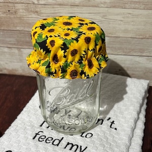 May include: A glass jar with a black and yellow sunflower fabric lid. The jar is labeled "Ball" and "Mason". The jar is sitting on a white towel with black embroidery that reads "I'm not to feed my sourdough"