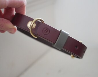 May include: A burgundy leather dog collar with a brass buckle and a grey leather accent. The collar has a small brass logo with the letters "&" inside a circle.