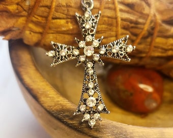 Könnte beinhalten: Ein silberfarbener Metall-Kreuzanhänger mit filigranen Details und eingelassenen klaren Strasssteinen. Das Kreuz hängt an einer dünnen silbernen Kette und zeigt ein symmetrisches Design mit spitz zulaufenden Enden und einem zentralen Stein.