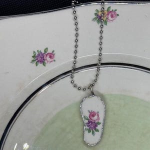 May include: A silver chain necklace with a pendant made from a piece of vintage china with a floral design. The pendant is shaped like a foot.