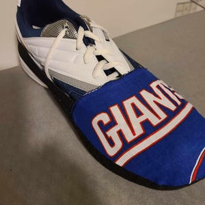 May include: A white and blue shoe with a black sole. The shoe has a blue fabric cover with the word "GIANTS" printed in white.
