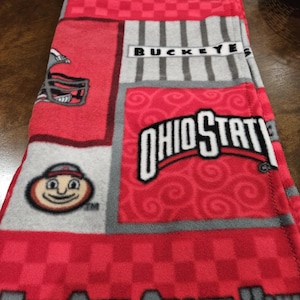 May include: A fleece blanket featuring a red, gray, and white patchwork design with The Ohio State University text, the Ohio State logo, a football helmet graphic, and the Brutus Buckeye mascot face. The fabric has a soft, textured surface.