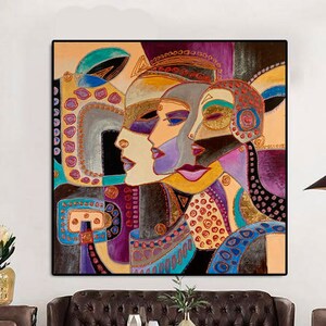 Op de afbeelding: Abstracte kunstprint met drie kleurrijke gezichten met geometrische patronen. De gezichten zijn in tinten goud, paars, blauw en rood. De print is ingelijst en hangt aan een witte muur.