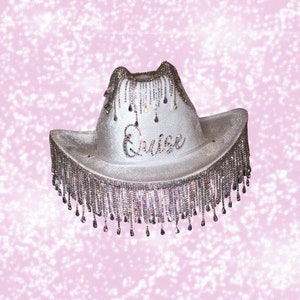 May include: A white cowboy hat with a fringe trim made of rhinestones. The hat has the word "Cruise" written on the front in rhinestones.