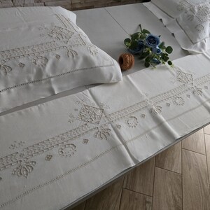 May include: White embroidered bedding set with a pillowcase, runner, and another pillowcase. The set features detailed floral and geometric patterns. A blue floral arrangement and a decorative wooden ball are placed on the bedding.
